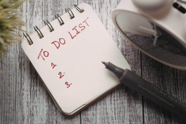 A small spiral notebook on a wooden surface with “To Do List” written at the top and numbers 1–3 below, next to a pen and a bedside clock.